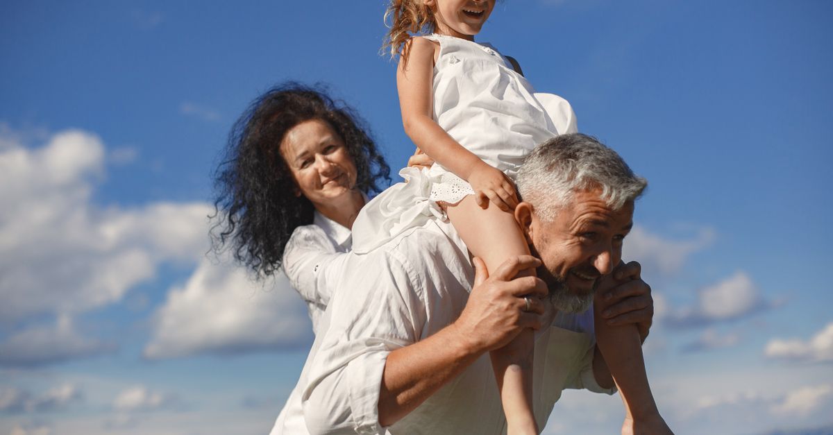 Grandparents playing with granddaughter outdoors under bright blue sky.