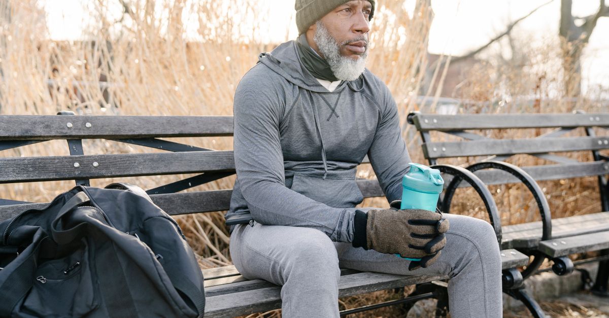 Senior African American bearded male in warm activewear sitting on wooden bench with sport bag and bottle of water in city park and looking away thoughtfully in daylight