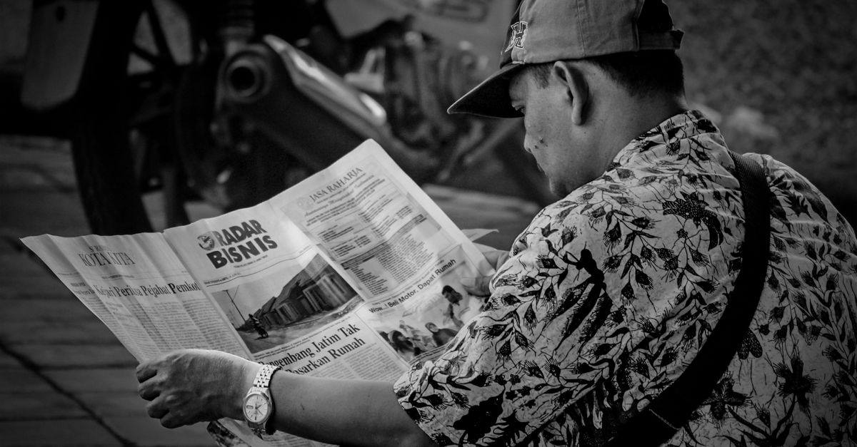 Black and white photo of a man reading a newspaper outdoors in Surabaya, Indonesia.