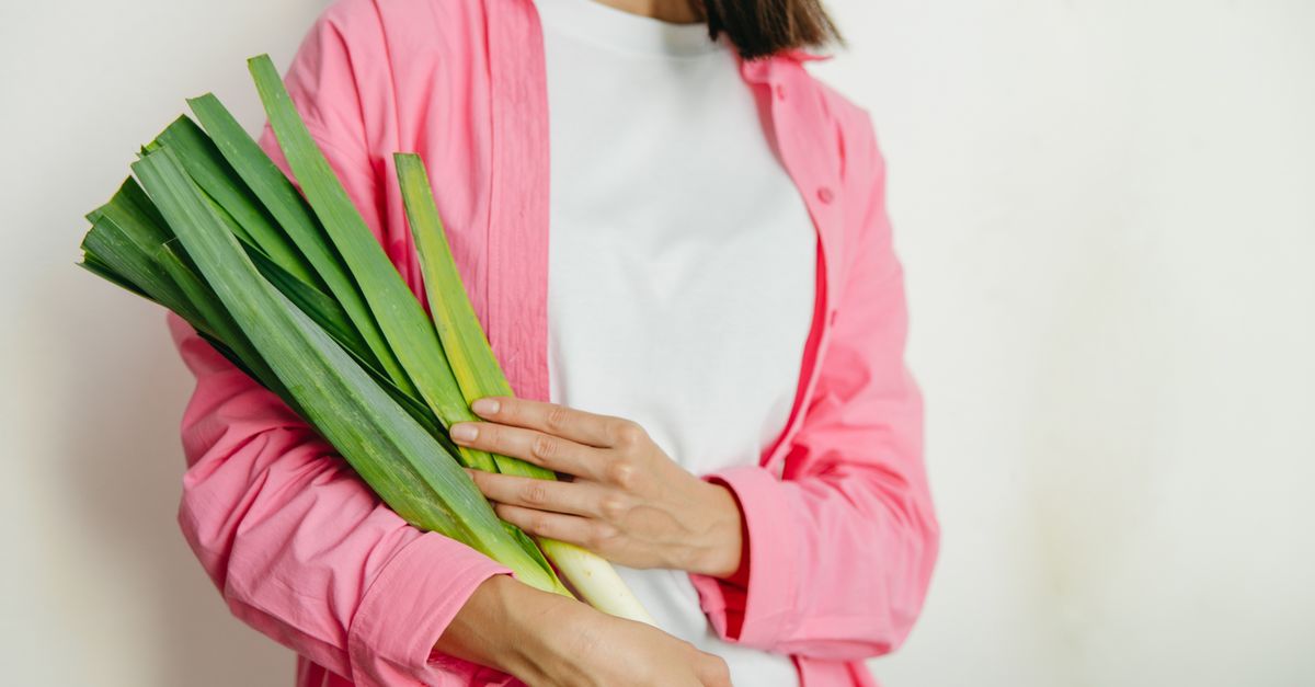 A woman in a pink jacket holding fresh spring onions against a white background.