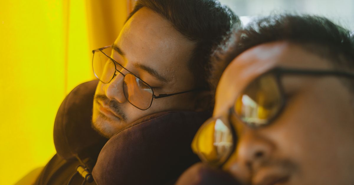 Two men wearing eyeglasses sleep on a bus, using neck pillows for comfort during the journey.