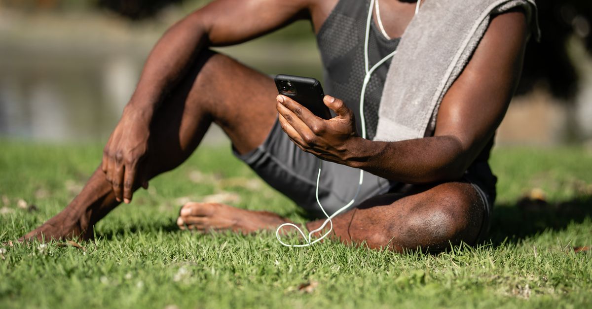 Adult man sitting on grass, using smartphone with earphones and towel. Outdoor relaxation.