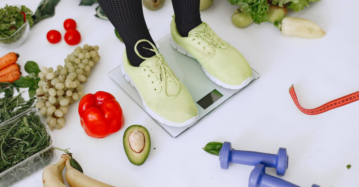 Person standing on scales surrounded by fresh produce and fitness equipment symbolizing a balanced lifestyle.