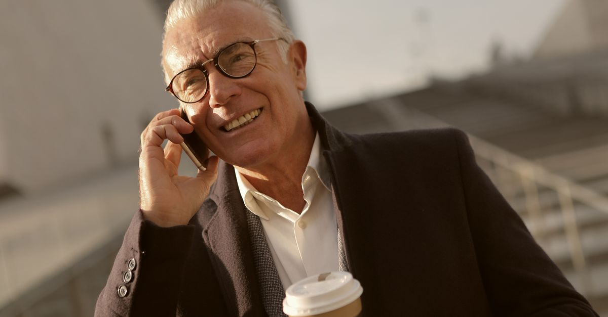 Elderly man smiling while talking on mobile phone with coffee in hand outdoors.