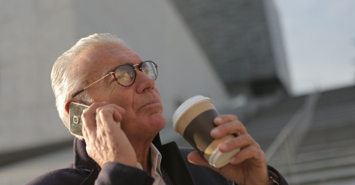 Elderly man in glasses using smartphone while holding a disposable coffee cup outdoors.