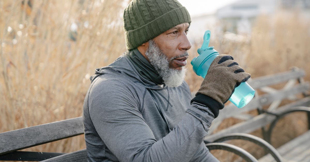 Side view of serious gray haired African American male in warm activewear sitting on wooden bench in park and drinking water while resting after training and looking away in daylight