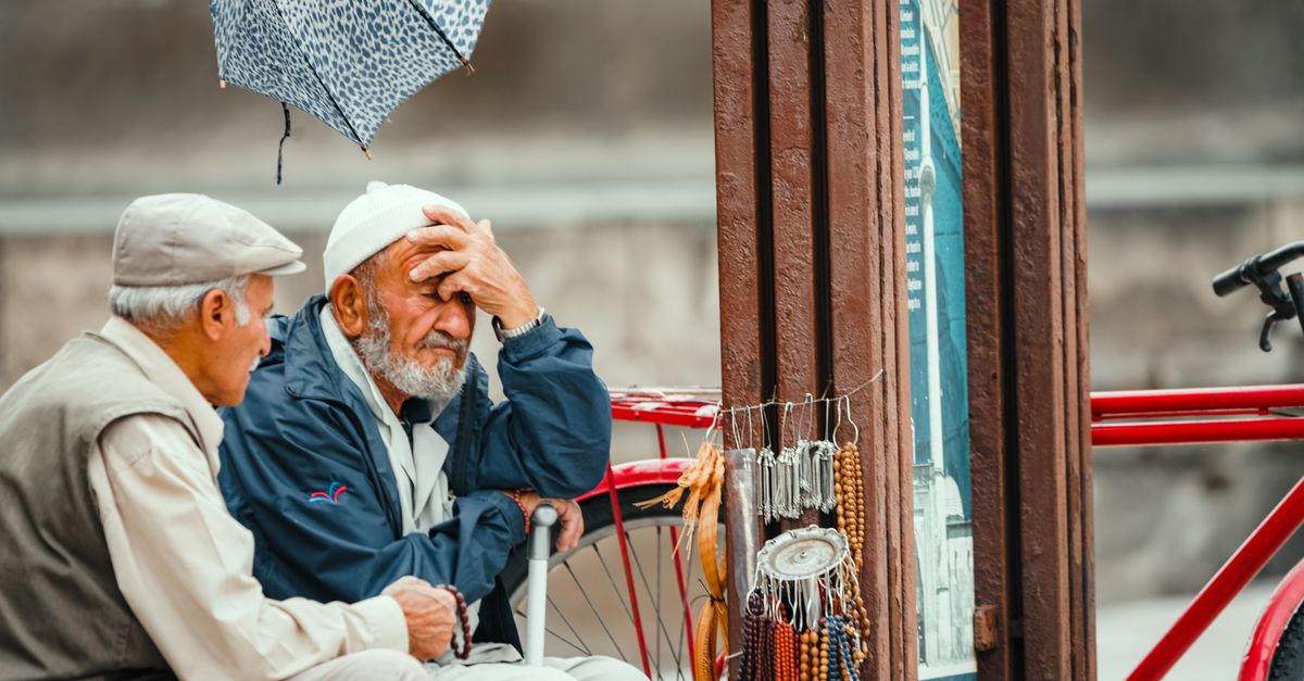 Two elderly men sit by a street vendor, engaged in conversation, with beads on display and a bicycle nearby.