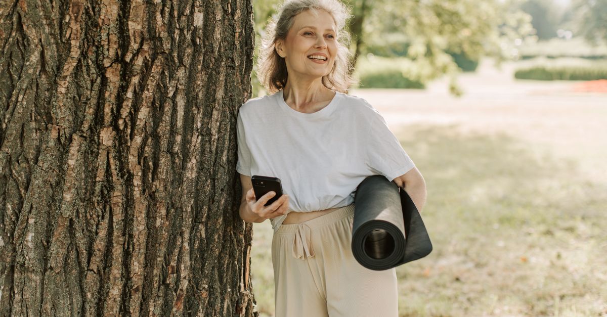 Happy senior woman with yoga mat and phone outdoors, enjoying nature and relaxation.