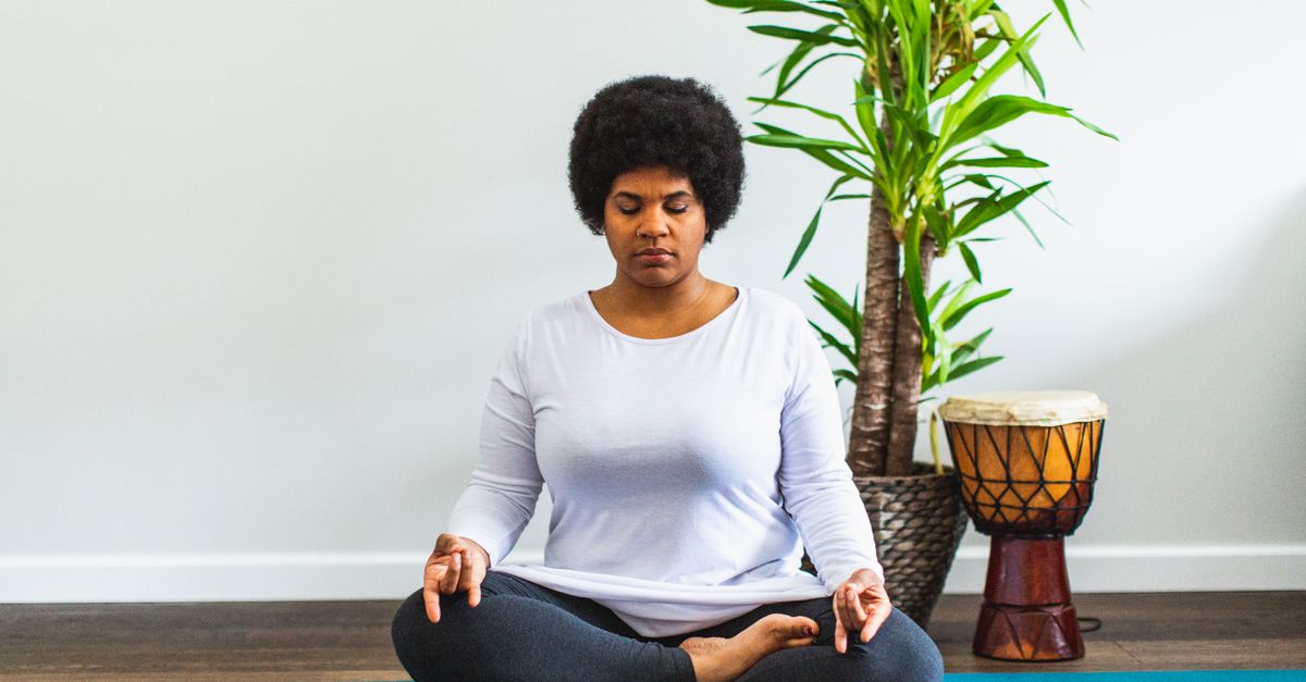 African American woman meditating indoors on a yoga mat with a plant and drum nearby.