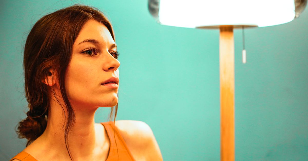 Side view of pensive female looking away while sitting near glowing lamp and green wall in modern apartment during weekend