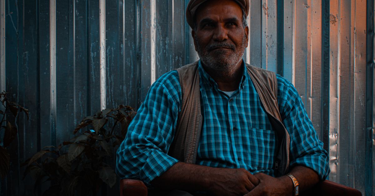 A contemplative man with facial hair sitting indoors, wearing a blue checkered shirt and flat cap.