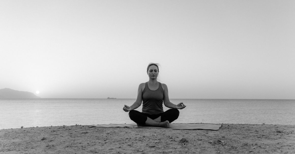 Black and white photo of a woman meditating by the sea during sunrise, creating a serene atmosphere.