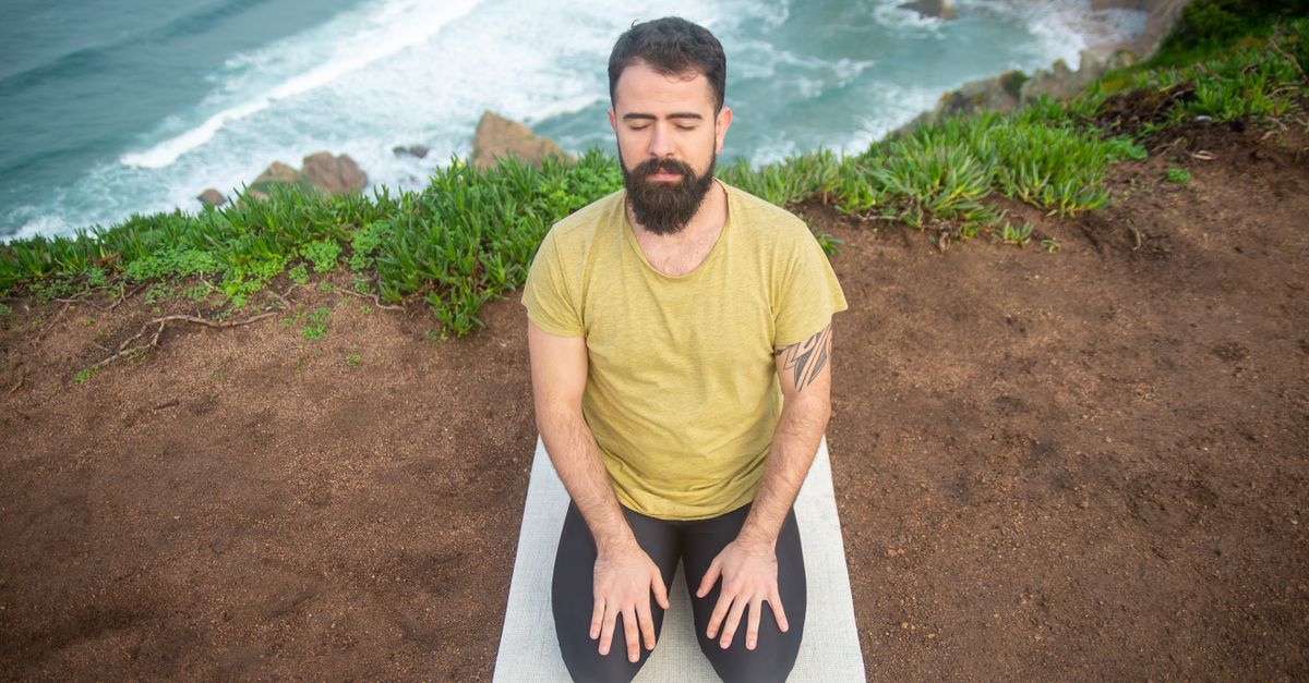 A man in a green shirt meditates by a cliff with ocean view, embracing tranquility.