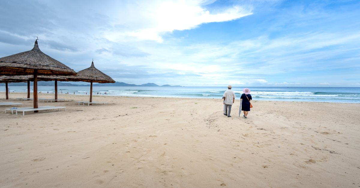 A peaceful scene of an elderly couple walking on a sandy beach under a partly cloudy sky.
