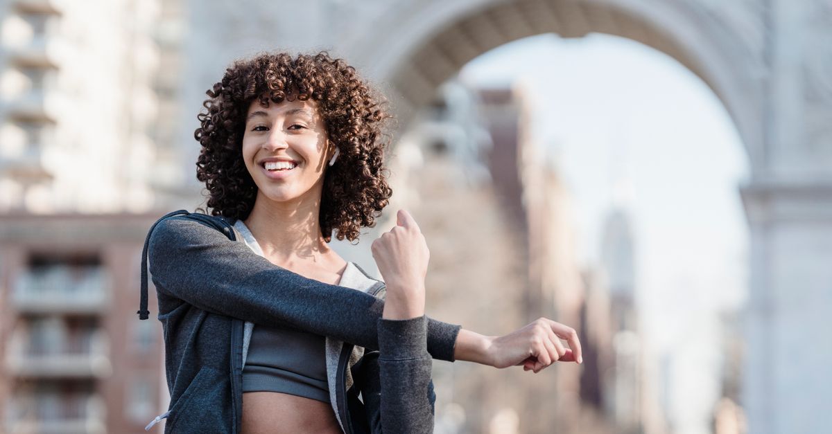 Delighted young ethnic female with Afro curls wearing sportswear stretching arms and listening to music in TWS earbuds while preparing for training