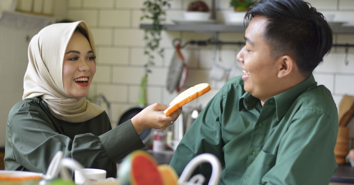 Smiling couple in a kitchen sharing bread, wearing green and a hijab, creating a cozy and loving atmosphere.