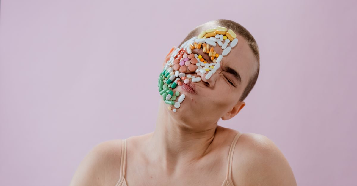 Conceptual portrait of a woman with pills on her face against a pink background, expressing health themes.