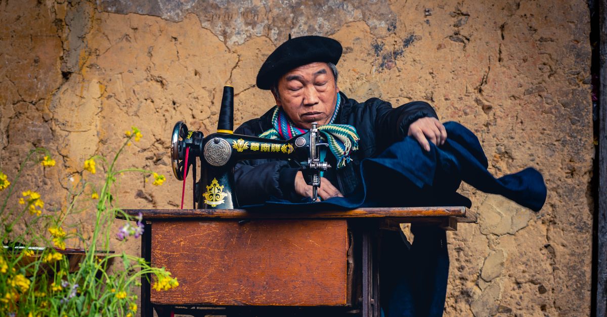 An Asian adult man sewing outdoors on a vintage sewing machine, wearing a colorful scarf and beret.