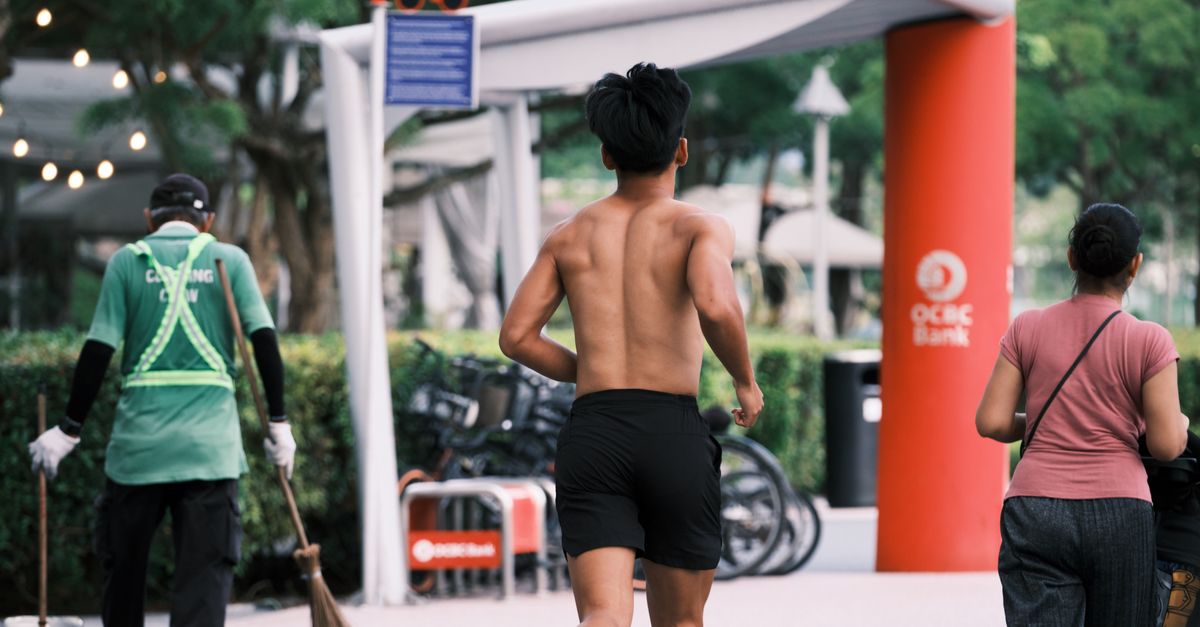 A shirtless man jogging on a sunny day in Singapore city amidst pedestrians and urban landscape.