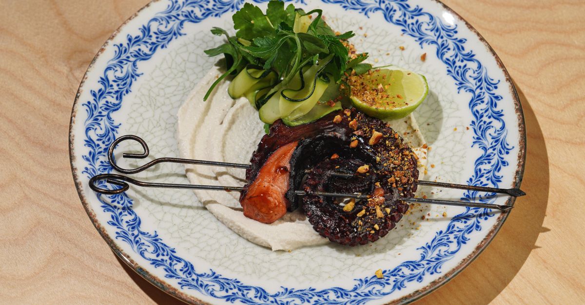 Gourmet octopus skewer with greens, lime, and spices on a decorative plate.