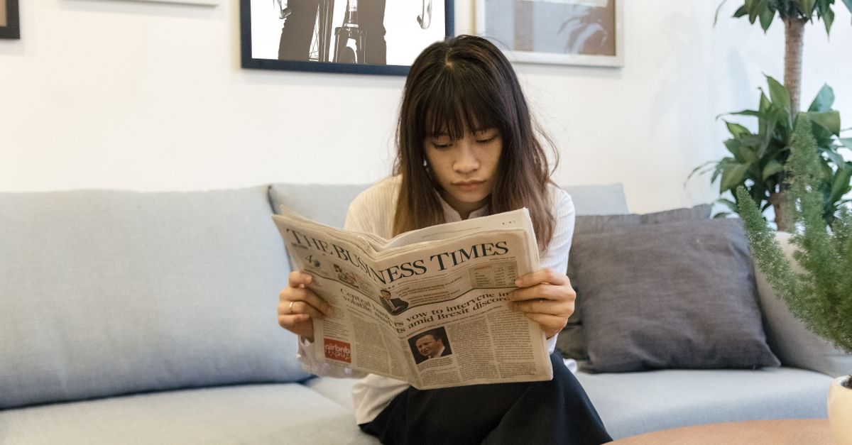 A young woman attentively reads a newspaper while sitting on a modern sofa indoors.