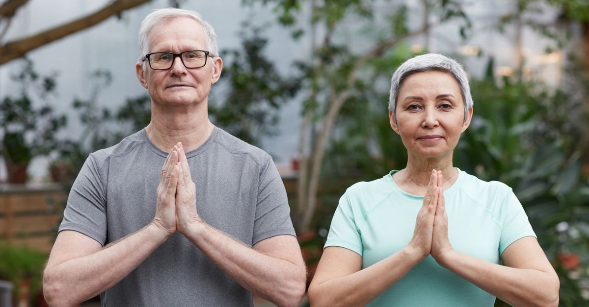 Senior couple performing yoga poses in a peaceful greenhouse setting.