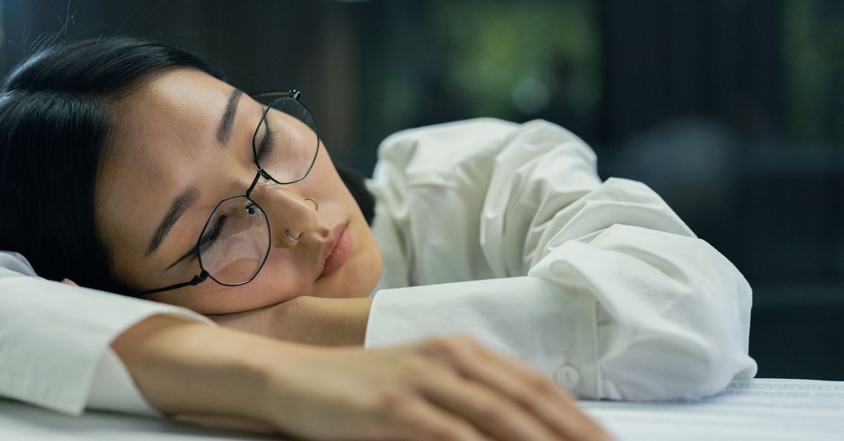 A tired woman with eyeglasses sleeps on a desk indoors, highlighting exhaustion.