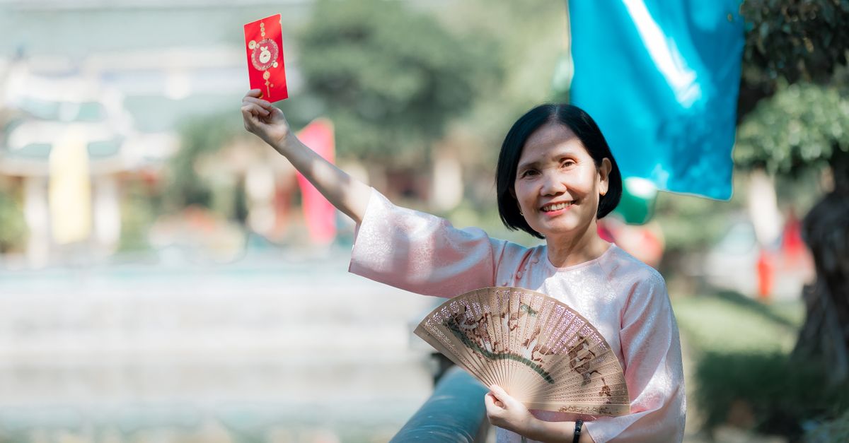 Smiling woman in traditional attire holding a red envelope and fan outdoors, symbolizing celebration.
