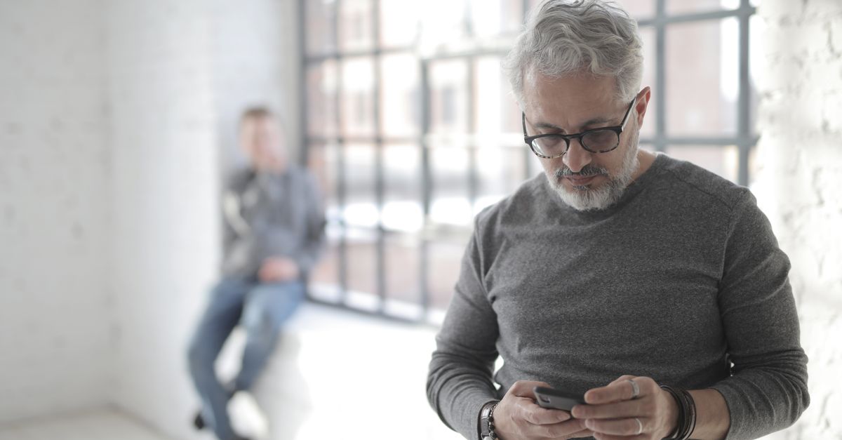 Focused mature male employee in eyeglasses and casual wear browsing internet on cellphone while standing near fenced window behind colleague in sunlight