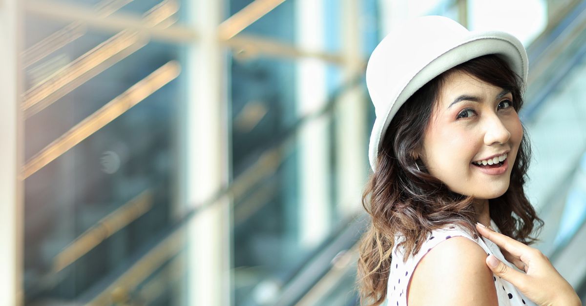 A cheerful young woman in a white hat smiles brightly by an escalator indoors.