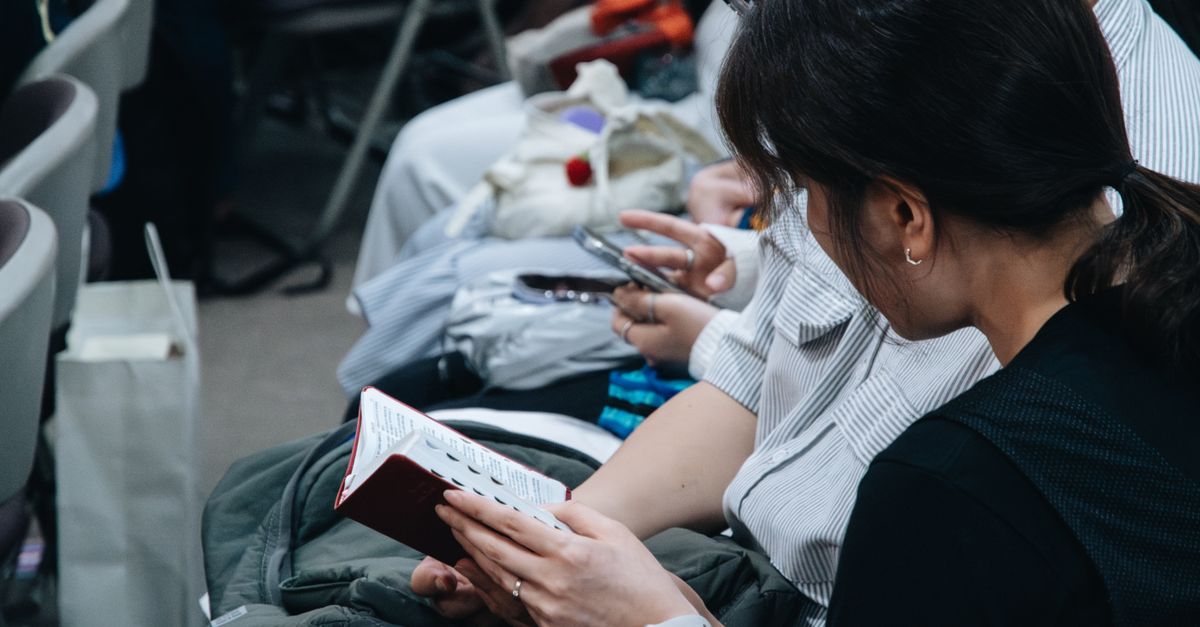 Close-up of people sitting in a crowd, reading a book and checking a smartphone.