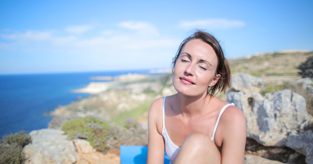 A woman relaxes with closed eyes on a scenic seaside cliff, embracing the warmth of summer.