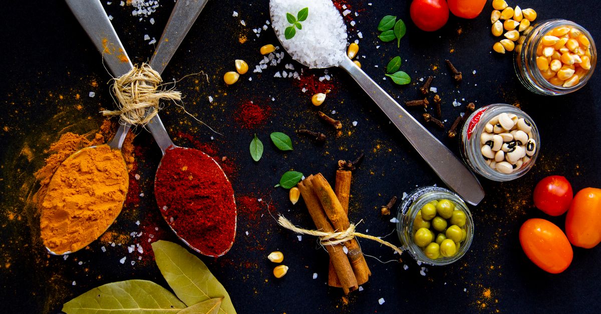 Top view of dry turmeric and paprika near bay leaves with cinnamon sticks and sea salt in spoons near jars with popcorn grains and olives with ripe colorful cherry tomatoes