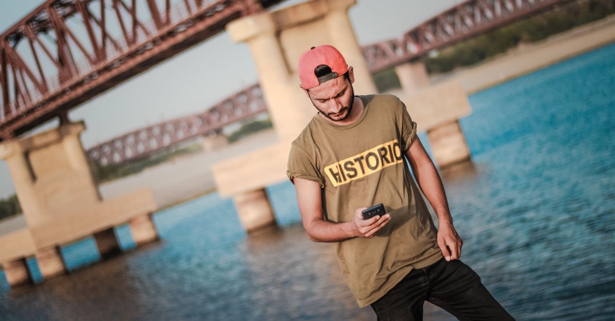 Casual young man with smartphone by a river bridge, enjoying outdoor summer leisure.