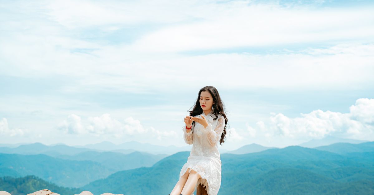 Full length of peaceful young ethnic lady with long dark hair in stylish dress relaxing on old house roof during trip in mountainous valley on sunny day