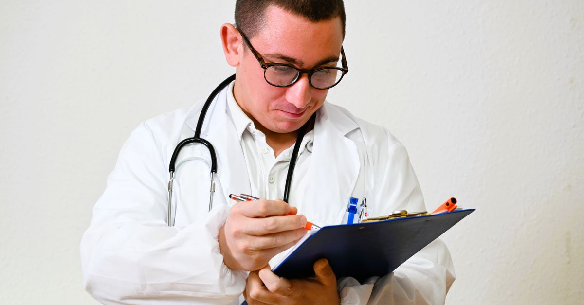 A male doctor in a white coat writes on a clipboard in a hospital setting, showcasing healthcare professionalism.