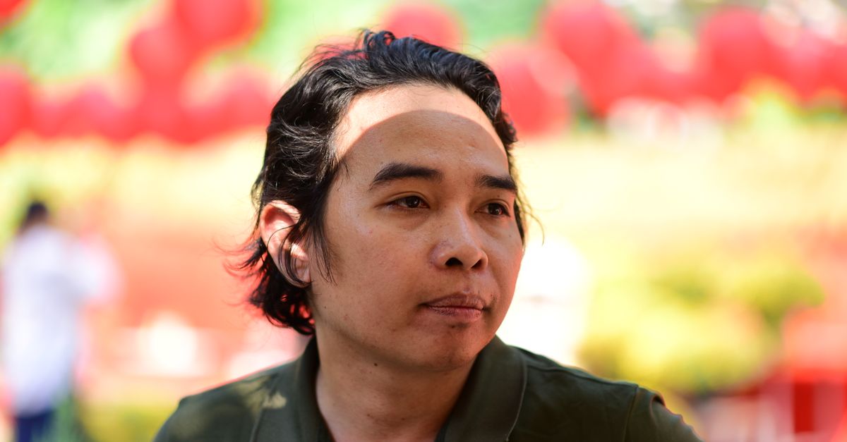 A thoughtful man stands outdoors surrounded by vibrant red balloons.