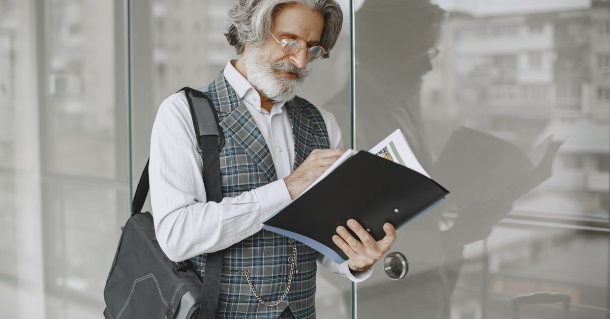 Elderly man in stylish office attire reading documents by glass windows.