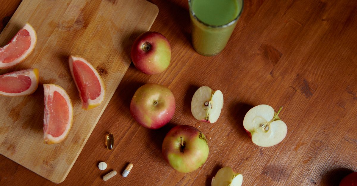 Top view of sliced grapefruit, apples, pills, and a green drink on a wooden table, emphasizing health.