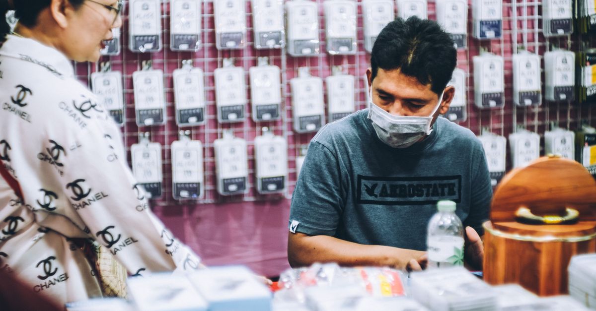 Ethnic man seller in protective mask sitting on counter in store with accessories next to Asian female buyer