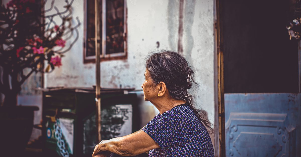 Elderly woman sitting by a doorway, gazing thoughtfully outdoors in a rustic setting.