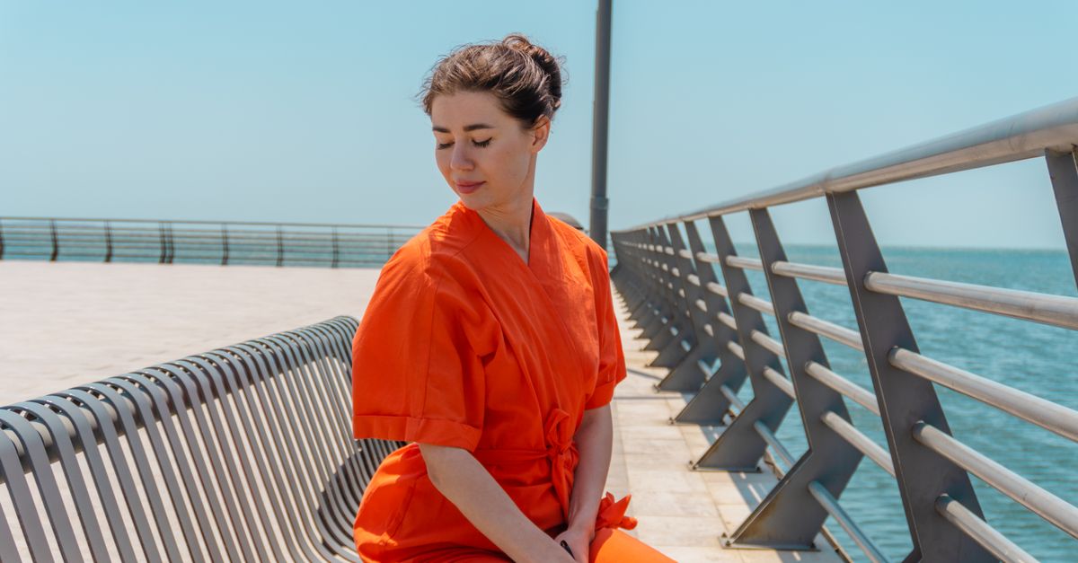 Woman in an orange outfit sitting on a bench along a seaside promenade.