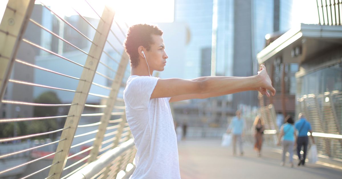 Focused African American man in earbuds and sportswear listening to music and stretching arms while training alone on street against blurred high rise buildings in downtown