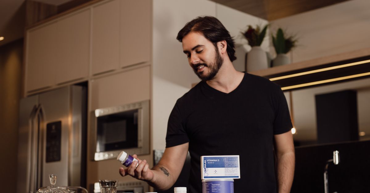 Man in a modern kitchen holding a vitamin supplement bottle, surrounded by health products.