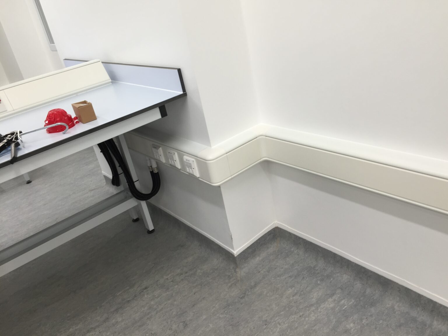 U-shaped PVC wiring duct mounted beneath a work surface on a white office wall, organizing electrical cables and sockets