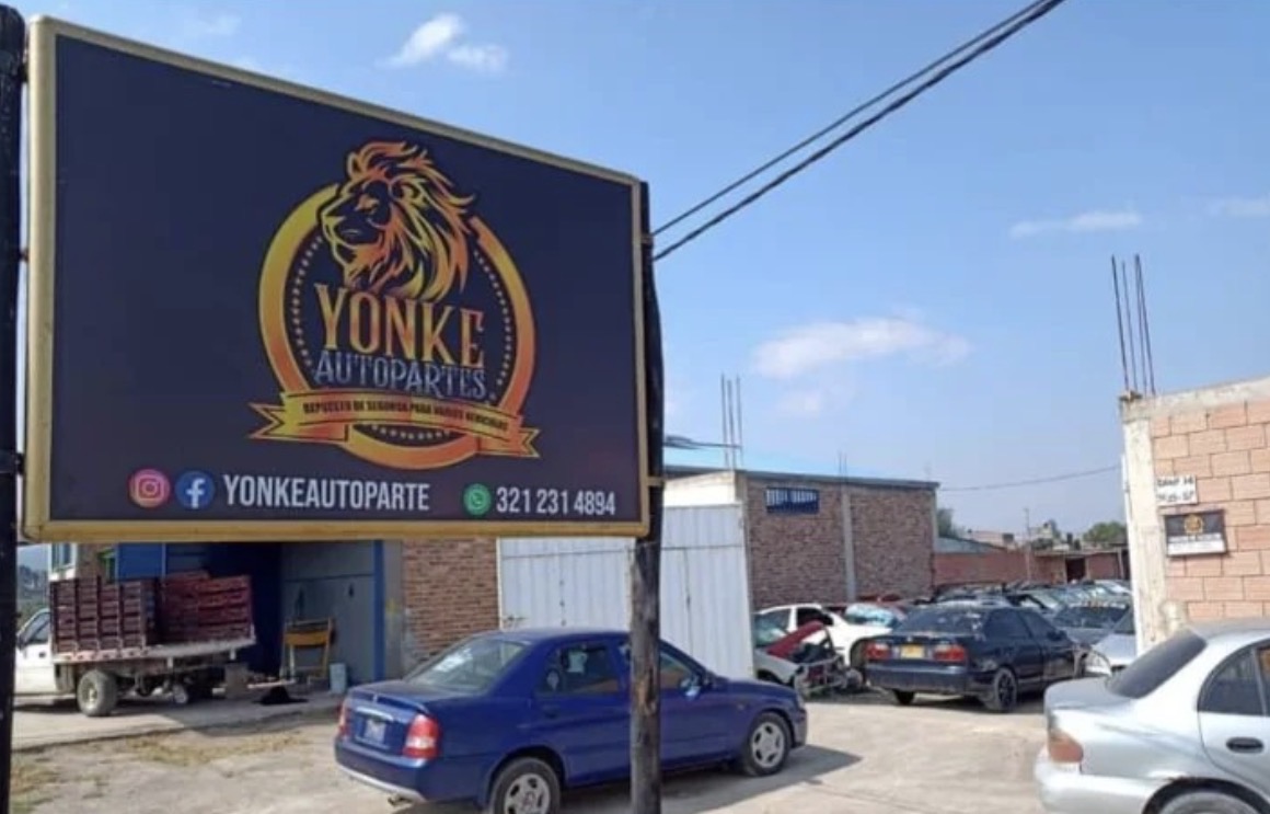 Aviso de YONKEAUTOPARTES en la entrada del patio con vehículos para desarme al fondo
