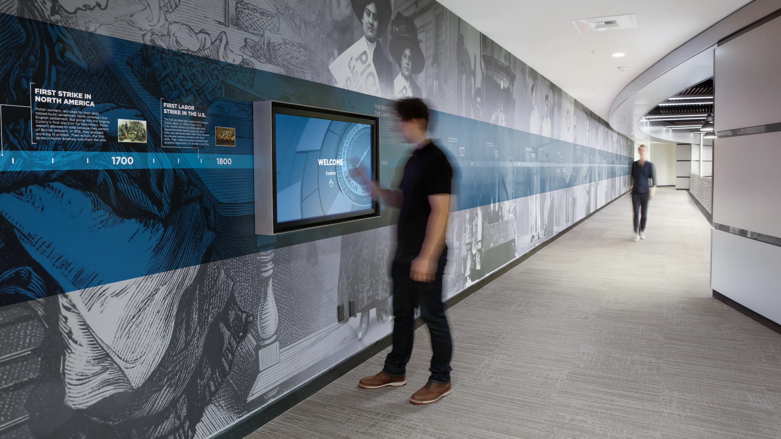 A man stands in front of a timeline wall graphic, interacting with a wall-mounted touchscreen interface