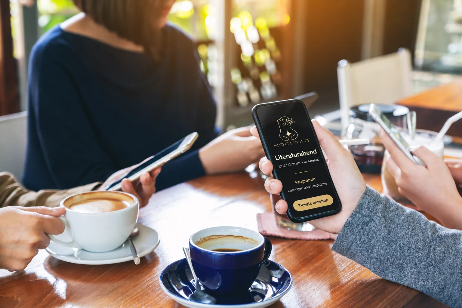 Menschen mit Smartphones am Cafétisch; auf dem vorderen Gerät ein Nocstar-Mockup zum Literaturabend.