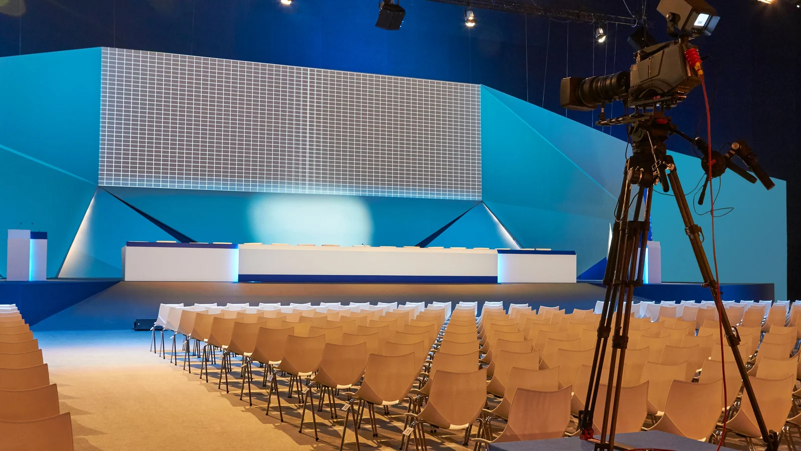 Leerer Konferenzsaal mit Stuhlreihen, Bühne und Kamera auf einem Stativ.