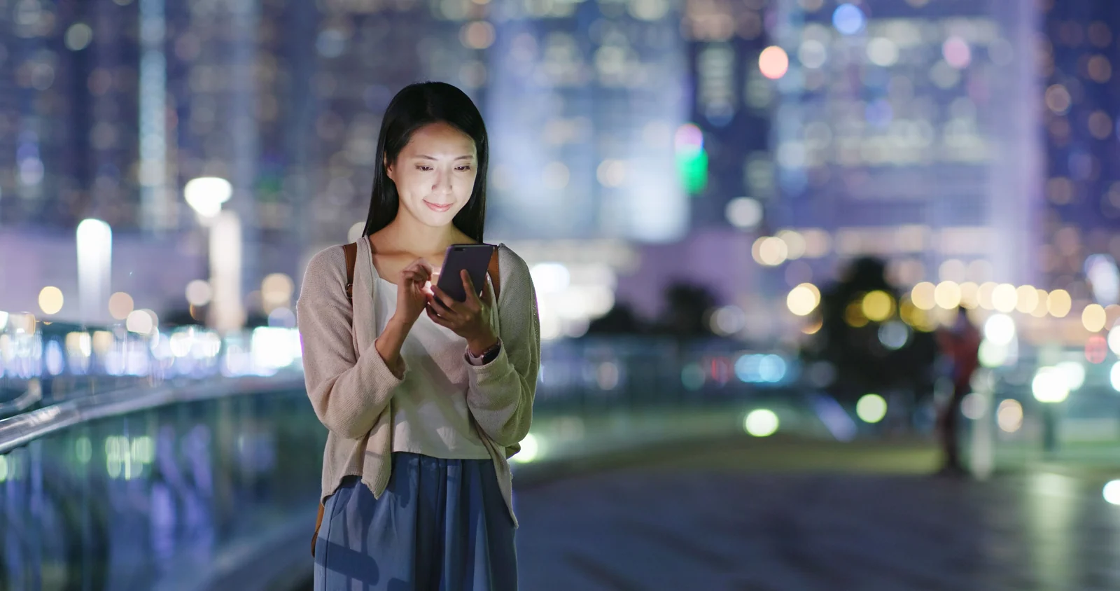 Eine Frau nutzt ein Smartphone vor unscharfen Stadtlichtern am Abend.